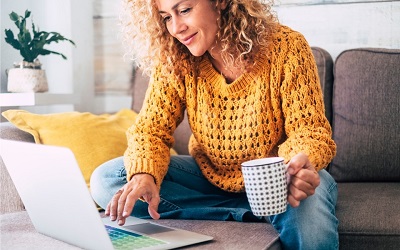 אישה עם חולצה בצבע חרדל ותלתלים מול מחשב נייד יושבת על ספה
